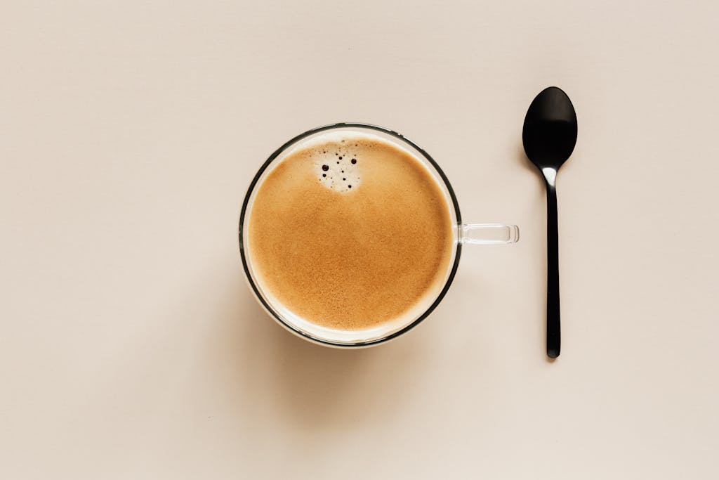 From above of clear glass cup with hot aromatic drink placed near black metal spoon on beige table for coffee break